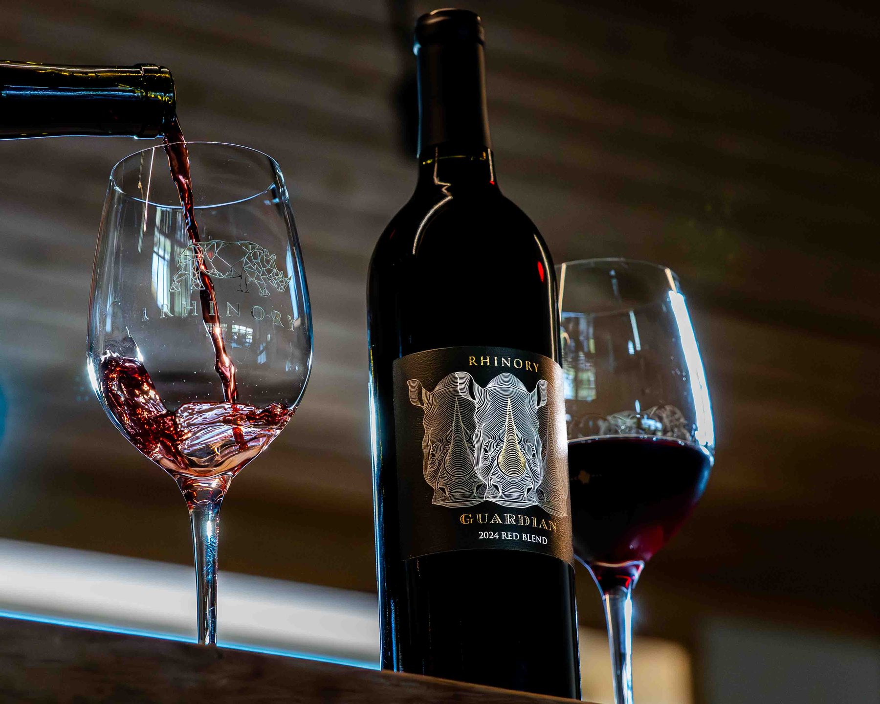 A red pour into an etched Rhinory glass beside a bottle of Rhinory Guardian red blend